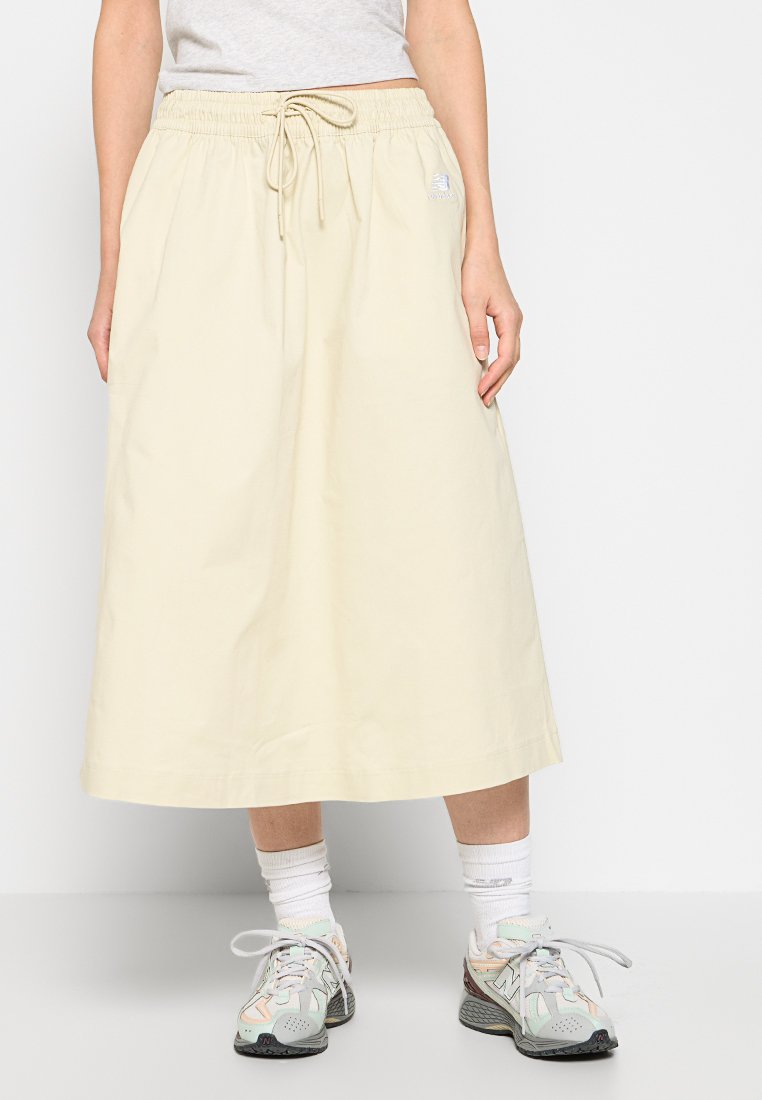 Falda beige clara con cintura elástica y cordón. Tiene un corte recto y cae por debajo de la rodilla. Se lleva con zapatillas multicolores.