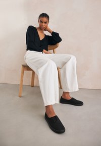 Sort langærmet bluse med butterfly, kombineret med hvide denim bukser med vide ben og sorte canvas slip-on sko. Lys baggrund, stol af træ.