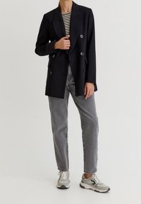 Femme portant un blazer noir croisé, une chemise rayée, un jean gris et des baskets blanc-gris, se tenant devant un fond uni.