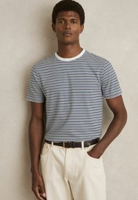 Tricou cu dungi în nuanțe de bleumarin și alb, cu guler rotund și mâneci scurte, asortat cu pantaloni de culoare crem și o curea închisă.