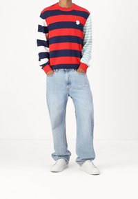 Pull tricot rayé avec des couleurs rouge, marine et aqua ; jean ample bleu clair, baskets blanches. Comprend des poignets côtelés et un détail de logo floral.