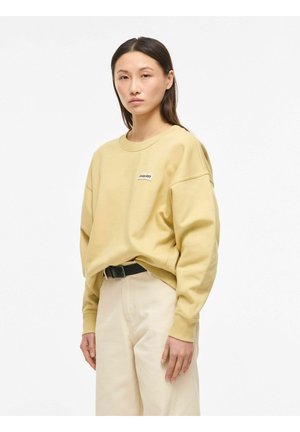 Fiatal nő oldalról áll, bézs színű, kerek nyakú pulóvert és krémszínű, magas derekú nadrágot visel fekete övvel, fehér háttér előtt.