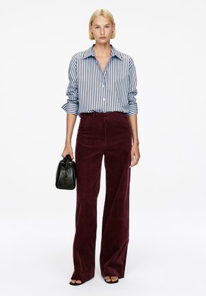 Frau mit kurzen blonden Haaren, die ein blau gestreiftes Hemd, burgunderfarbene Cordhosen, schwarze Sandalen trägt und eine schwarze Handtasche hält.