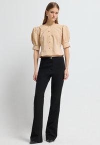 Vrouw in een beige blouse met pofmouwen en knopen, en zwarte hoge taille flare broek, tegen een effen lichte achtergrond.