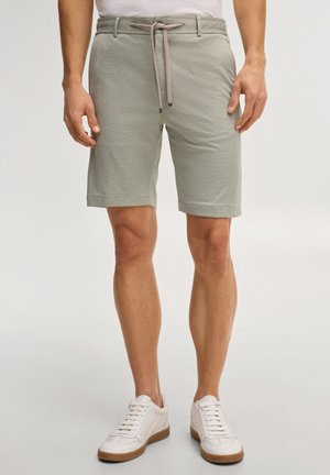 Mężczyzna noszący jasnoszortki ze strukturą i sznurkiem oraz białe trampki, stojący na jednolitym tle.