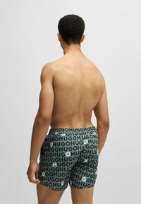 Green swim shorts featuring a repeating "HUGO" logo pattern in varying shades. Elastic waistband, loose fit, and smooth fabric texture.