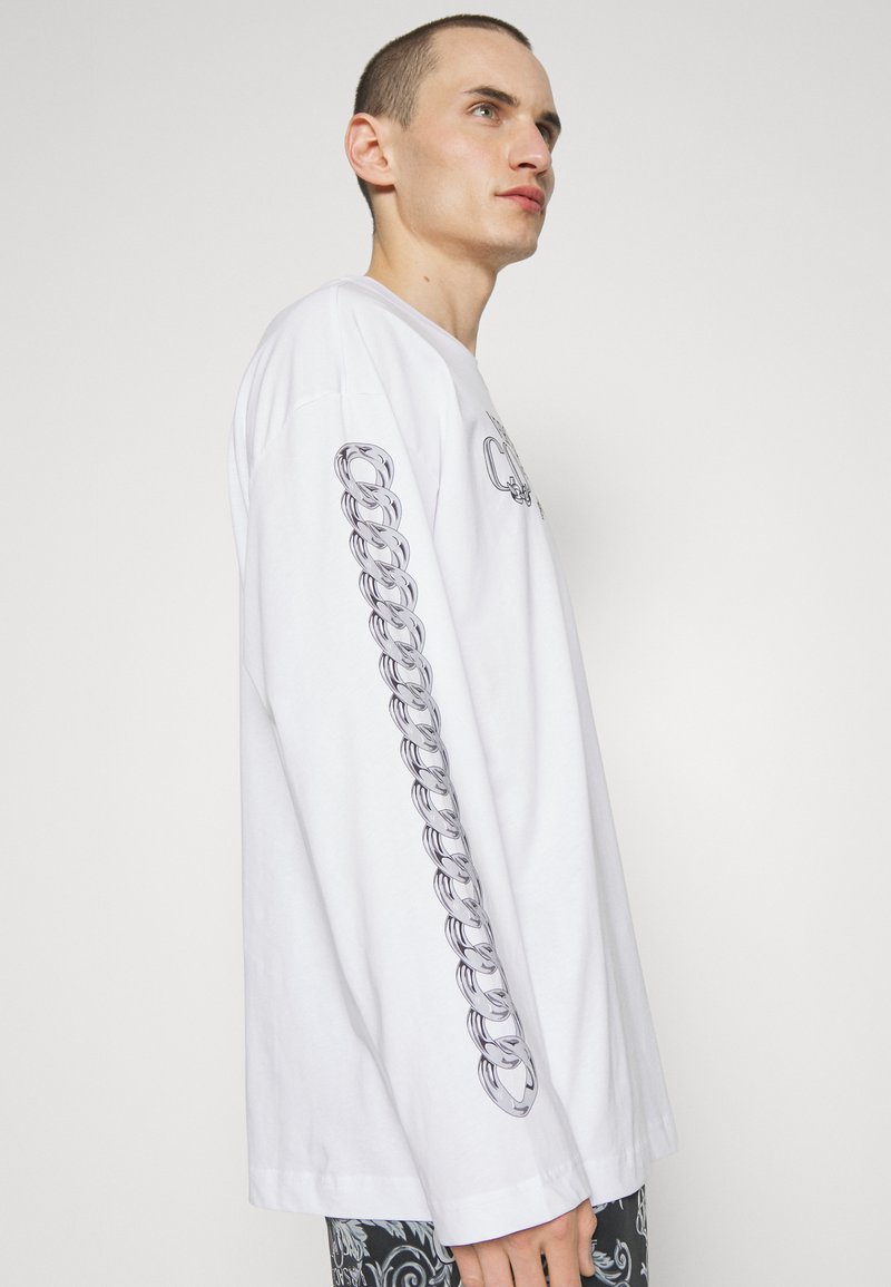 Camiseta blanca de manga larga con un diseño gráfico de eslabones en la manga izquierda. Tejido suave con un ajuste relajado y cuello redondo.