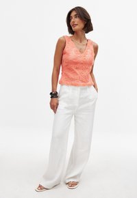 Peach floral-patterned sleeveless top with a V-neck, paired with loose white linen pants, and sandals. Accents include bracelets on one wrist.