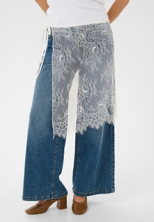 Person trägt eine weit geschnittene blaue Jeans, braune Schuhe mit Absatz, ein weißes Oberteil und eine weiße Spitzen-Schürze mit Blumenmuster, die in der Taille gebunden ist.