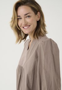 Top lejer, de culoare taupe, realizat dintr-un material ușor, cu decolteu în V, detalii pliate și mâneci moi, vaporose.
