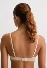 Femme avec des cheveux auburn ondulés attachés en une queue de cheval, portant un soutien-gorge beige, tournée vers un mur blanc uni, vue de dos.