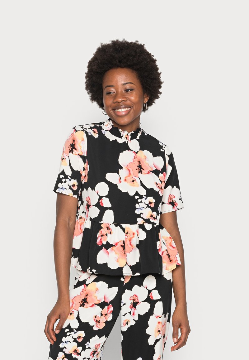 Top peplum floral com fundo preto, grandes flores brancas e rosas, mangas curtas e colarinho com babados. Combinado com calças correspondentes.