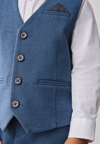 Chaleco azul con estampado, que presenta una tela texturada, cinco botones y un pañuelo en el bolsillo con un patrón de estrellas. Combinado con una camisa blanca texturada.