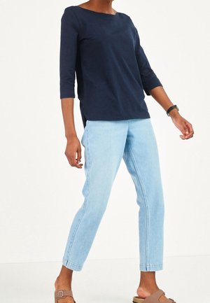 Personne portant un haut bleu marine à manches trois-quarts, un jean bleu clair longueur cheville et des sandales beige à bout ouvert avec une lanière à boucle.