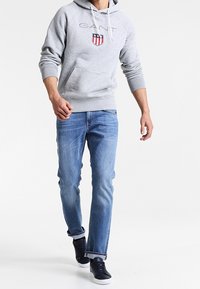 Felpa grigia con coulisse bianche e logo ricamato, abbinata a jeans azzurri chiari e scarpe da ginnastica scure. Abbigliamento casual per un look rilassato.