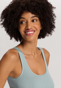 Top lungo azzurro realizzato in tessuto morbido. Presenta spalline larghe e un collo a barchetta. Il modello ha i capelli ricci e un trucco dai toni naturali.