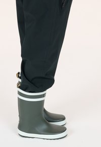 Bottes de pluie en caoutchouc vertes avec une semelle plate, des rayures blanches en haut et en bas, et une surface texturée, portées avec un pantalon imperméable noir.