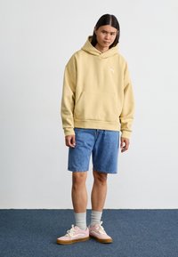 Felpa gialla con cappuccio e tasca frontale, abbinata a pantaloni shorts di denim blu. La calzatura presenta sneakers rosa con accenti bianchi e suole beige.