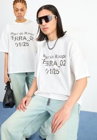 White oversized t-shirts with metallic text, paired with light blue, loose-fitting jeans. Accessories include a black bag and sunglasses.