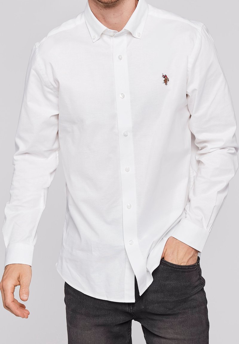 Homme portant une chemise blanche à manches longues avec un petit logo cheval brodé sur la poitrine et un pantalon noir.