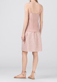 Žena v růžovém tílku a krátké růžové květované sukni, stojící bosky na jednoduchém světle šedém pozadí.