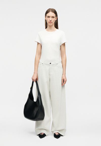 Naine, kes kannab valget t-särki ja laiade säärtega valgeid pükse, hoides vasakus käes suurt musta nahast käekotti, seisab tavalise tausta ees.