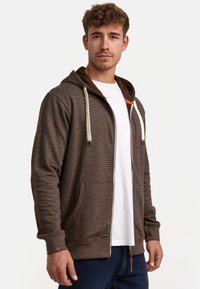 Brown zip-up hoodie with a textured pattern, contrasting inner hood, cream drawstrings, and side pockets. Fitted design, casual style.