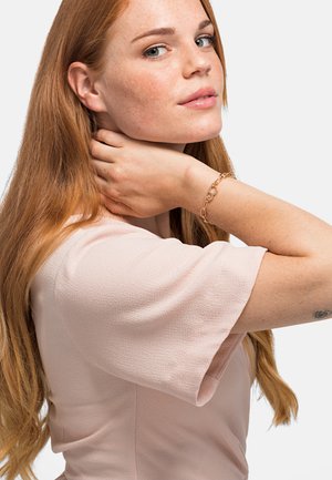 Blusa corta a maniche corte di colore rosa chiaro con tessuto testurizzato. Il modello indossa un bracciale di catena dorata e ha lunghi capelli rossi ondulati. Sfondo neutro.