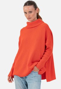 Pull orange oversize avec un col haut côtelé, des poignets de manches côtelés et une coupe décontractée. Porté avec un jean bleu clair.