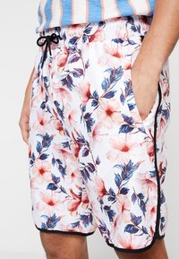 Shorts de bain fleuris en tissu blanc avec des motifs d'hibiscus roses et bleus. Dotés d'une taille élastique et de poches latérales avec un liseré noir.