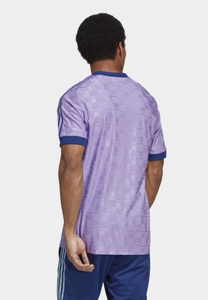 Homme portant une chemise à manches courtes lavande avec un motif à carreaux et une bordure bleu marine, associée à un pantalon bleu marine avec des rayures bleu clair.