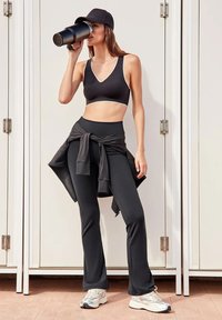 Zwarte sportbeha met witte afwerking, hoge zwarte leggings en een zwarte jas geknoopt om de taille. Grijze sneakers. Neutrale achtergrond.