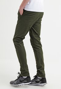 Olivgröna byxor med slim fit, tillverkade av textilmaterial. Bärs med svarta sportskor med ett gråmönster och snörning.