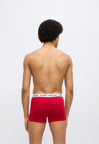 Κόκκινα ανδρικά εσώρουχα boxer με μαύρη ζώνη μέσης που φέρει το "TOMMY HILFIGER" σε λευκό. Το ύφασμα φαίνεται λείο, εφαρμοστός σχεδιασμός. Πλάγια όψη από πίσω.