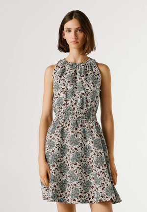 Frau trägt ärmelloses, knielanges Kleid mit gerafftem Ausschnitt und Taille, mit grün-braunem Blumenmuster auf weißem Hintergrund.