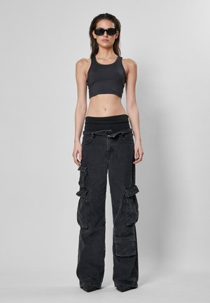 Svart avkortet tank top sammen med løse, baggy svarte bukser i cargo-stil med flere lommer og en teksturert finish. Svarte solbriller fullfører looken.