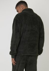 Man die zich omdraait met een donkergroen pluche fleece jas en bijpassende broek, tegen een effen lichte achtergrond.