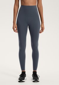 Höga, figurnäraleggings i mörkgrått stretchigt tyg, med slät yta och rena sömmar, kombineras med svarta sportskor.