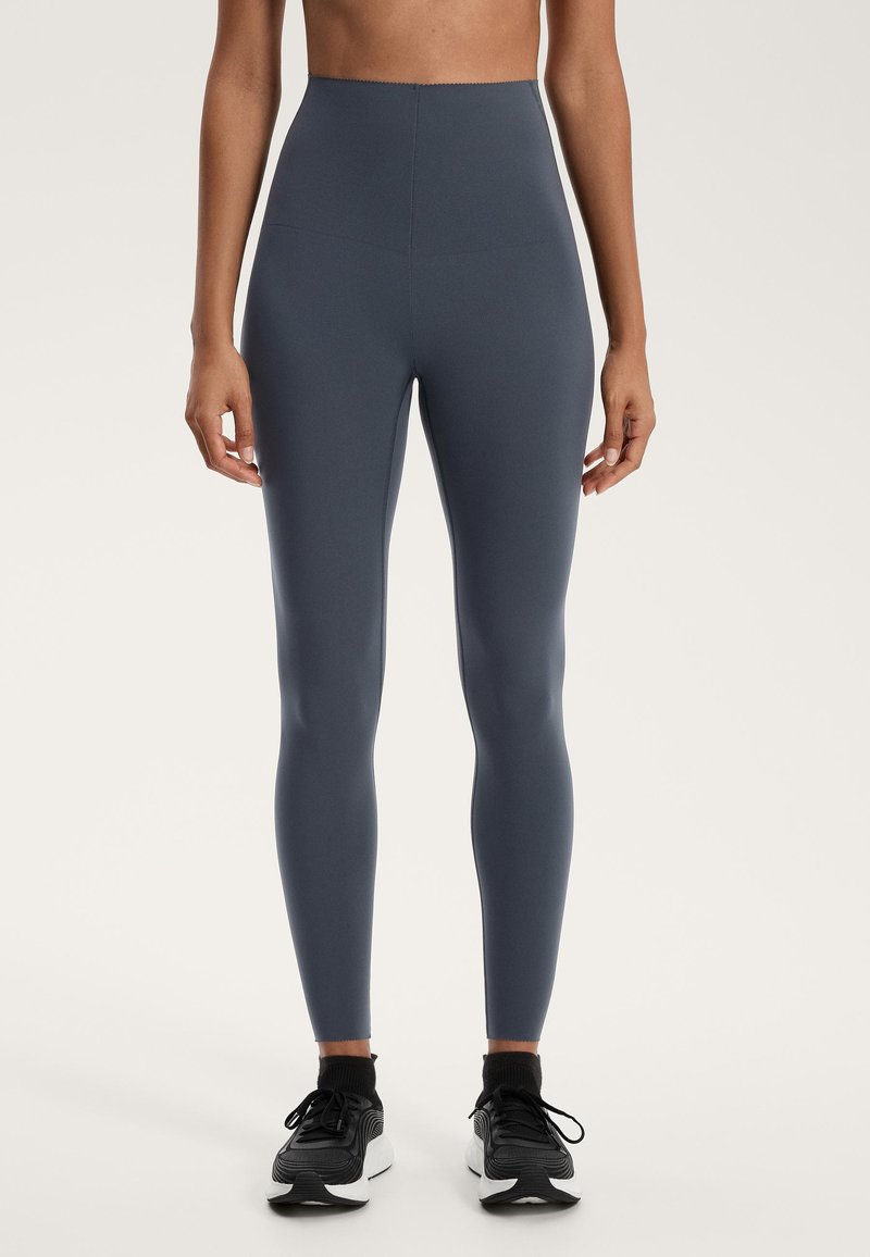 Höga, figurnäraleggings i mörkgrått stretchigt tyg, med slät yta och rena sömmar, kombineras med svarta sportskor.