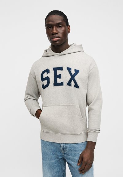 Pilka džemperio su gobtuvu, tamsiai mėlynomis raidėmis "SEX", su priekinė kišene ir siaurėjančiais rankogaliais, derinama su šviesiai mėlynais džinsais.