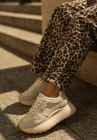 Białe sneakersy z teksturowanymi panelami i masywną podeszwą. W połączeniu z spodniami w leopardzi wzór. Plenerowe tło z schodami w tle.