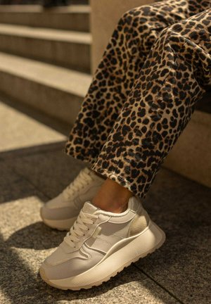Białe sneakersy z teksturowanymi panelami i masywną podeszwą. W połączeniu z spodniami w leopardzi wzór. Plenerowe tło z schodami w tle.