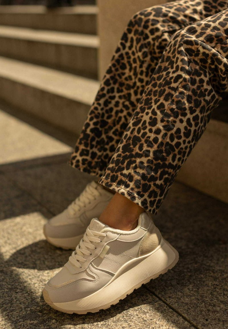 Białe sneakersy z teksturowanymi panelami i masywną podeszwą. W połączeniu z spodniami w leopardzi wzór. Plenerowe tło z schodami w tle.