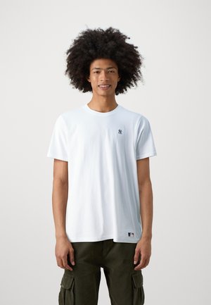 Maglietta bianca in cotone a maniche corte con un piccolo logo nero sul petto. Indossata con pantaloni cargo verde oliva. Tessuto liscio, vestibilità classica.
