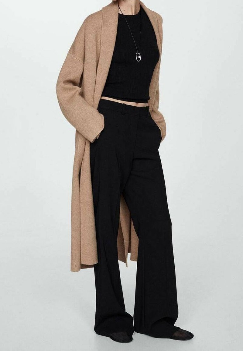 Personne portant un long manteau beige par-dessus un haut court noir, un pantalon noir à jambes larges et un collier avec un pendentif rond.