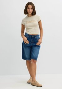 Camiseta corta beige con texto impreso, combinada con shorts de mezclilla azul de corte bermuda con cintura alta y bolsillos frontales, complementada con zapatos slip-on de color tostado.