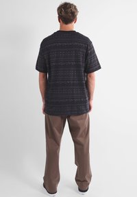 Camicetta nera a maniche corte con un design geometrico, abbinata a pantaloni marroni larghi. La camicetta ha un colletto a coste ed è realizzata in tessuto intrecciato.