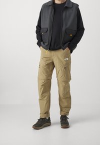 Grå fleeceväst över en svart långärmad tröja, kombinerat med lätta beige cargobyxor med dragkedjefickor och uppvikta fållar. M örka skor.