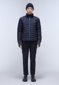 Námořnický prošívaný kabát s vodorovným švem, modrými zipovými detaily a pletenou čepicí. Nošen s tmavými kalhotami a černými botami.