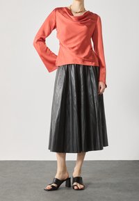 Top corallo con collo a cowl e maniche lunghe; gonna midi plissettata nera con texture lucida; sandali neri con tacco a blocco e cinturini aperti.